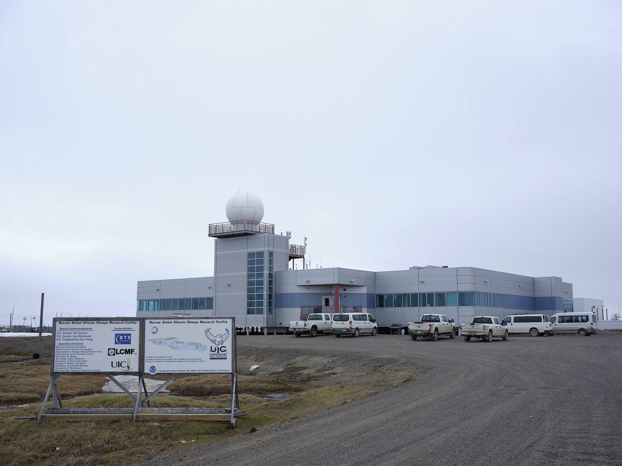 The Barrow Arctic Research Center, or BARC. A grey building with large glass windows with trucks parked in front